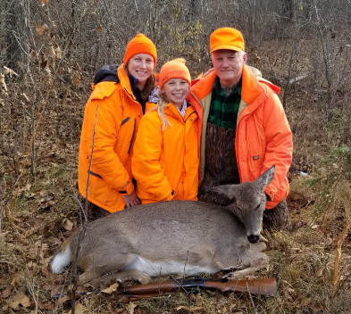 two adults and a child wearing blaze orange with a dead deer
