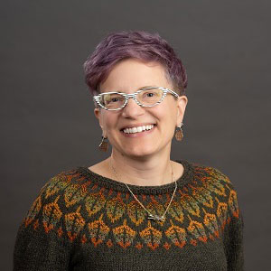 A woman with light purple short hair. Wearing glasses and a sweater