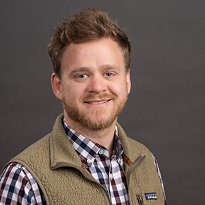 A young man with short hair and a beard wearing a plaid shirt and a vest