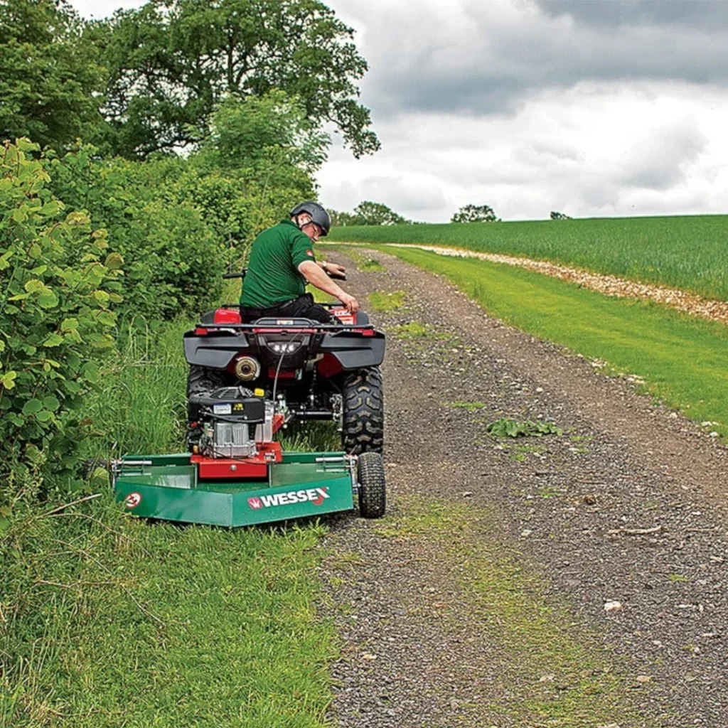 A person operating an ATV pull-behind mower along the edge of a forest