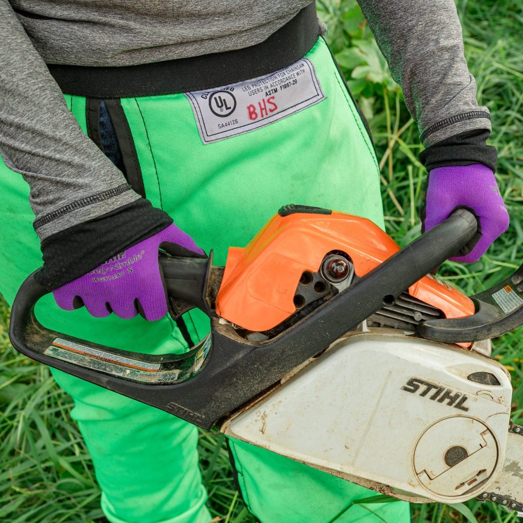 a person wearing gloves and chaps holding a chainsaw