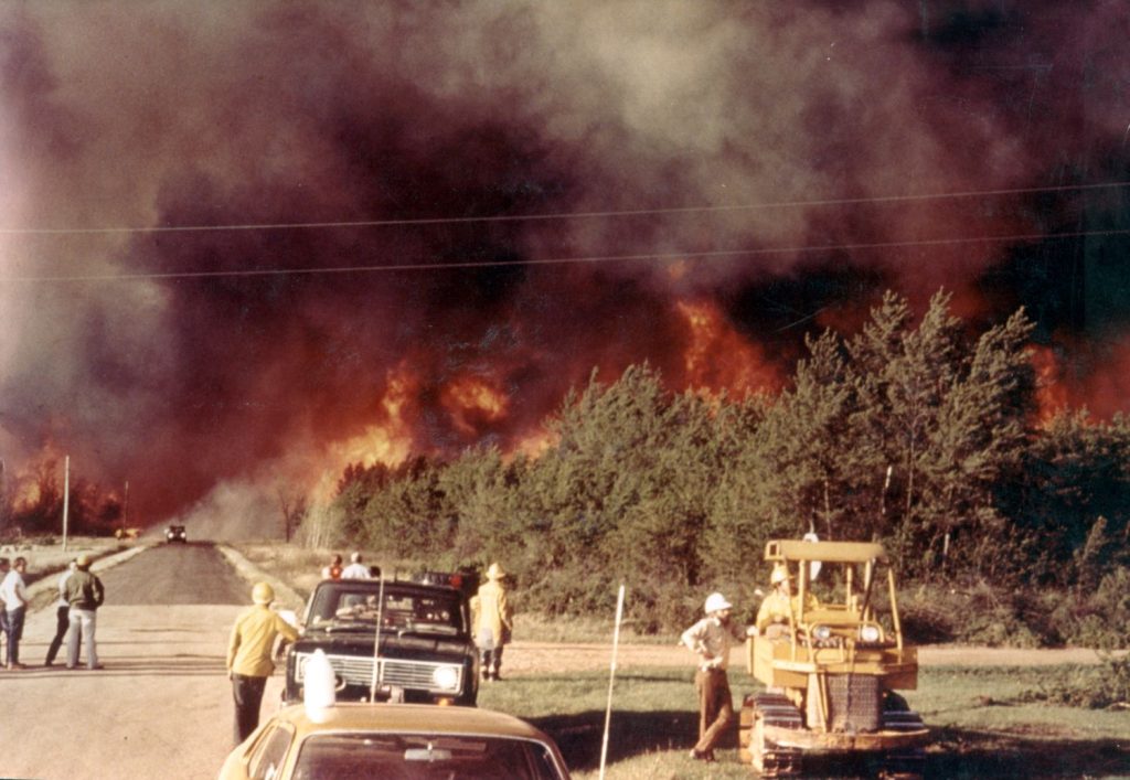People standing next to vehicles looking at a wildfire in the distance