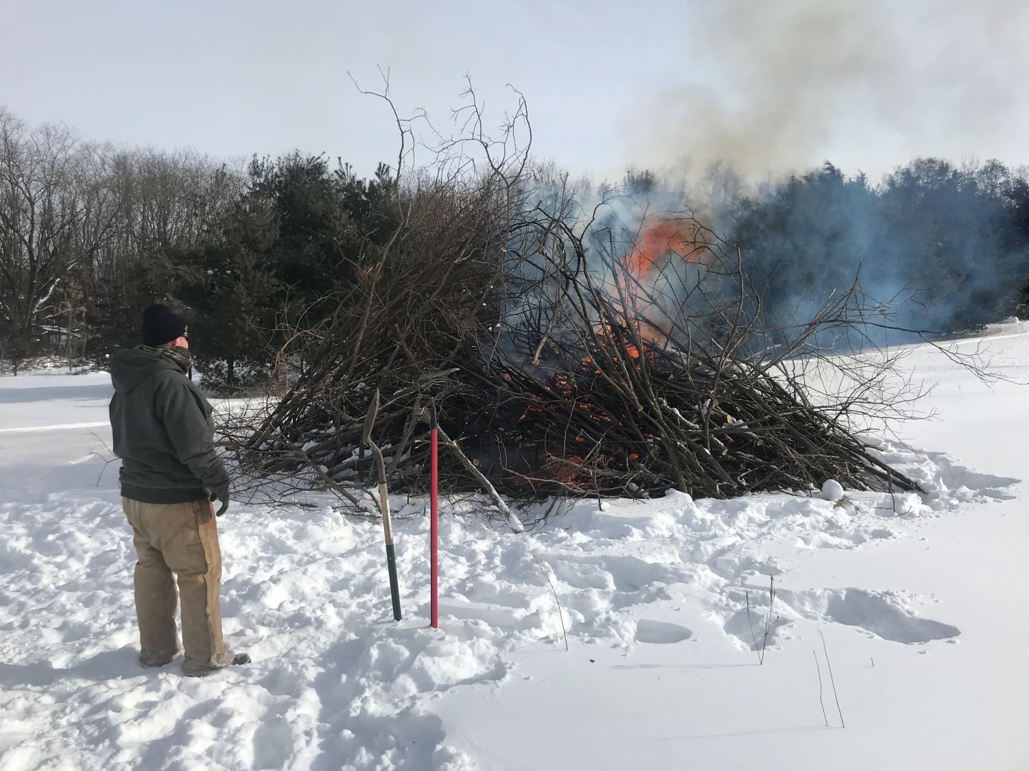 A person watching a controlled burn of a brush pile with snow-covered ground
