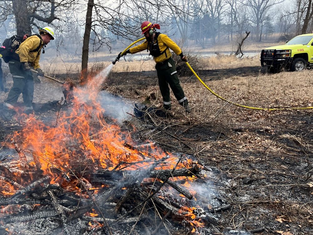 Two people in firefighting gear pouring water from a hose on a small fire
